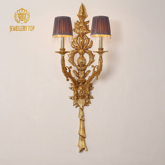 The ornate JEWELLERY TOP brass wall lamp installed on a white wall, showcasing its full Baroque grandeur and brand logo.