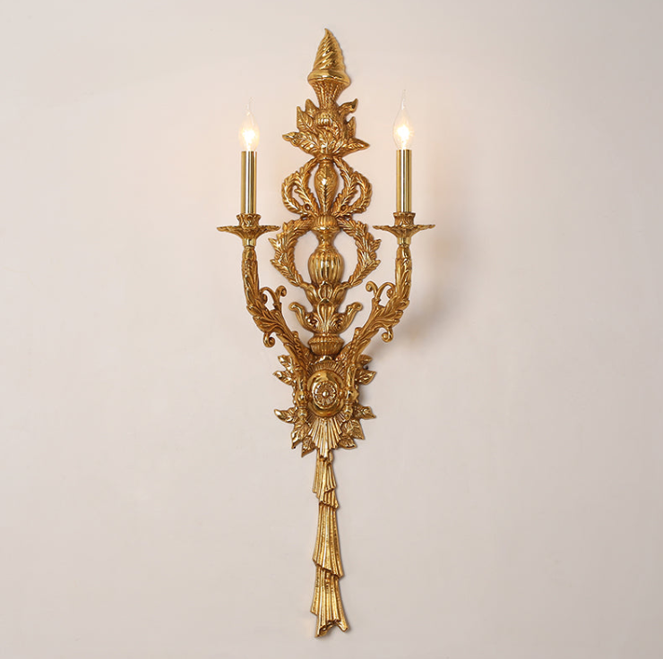 Front view of the antique brass wall lamp with intricate floral carvings and glowing candle-style lights on a white wall.