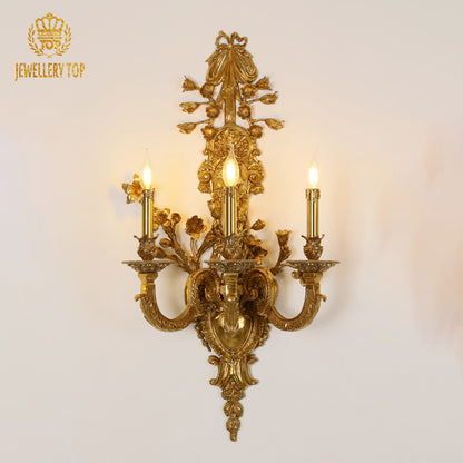 Front view of the JEWELLERY TOP Royal Noeud Lumière brass wall sconce with ornate bow and three lights on a white wall.