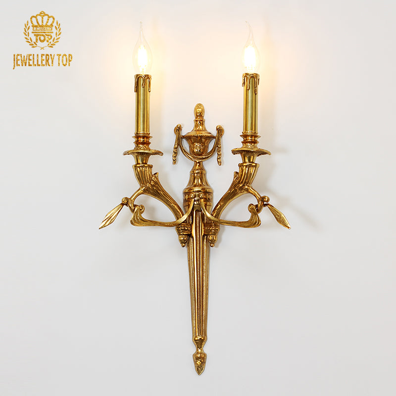 JEWELLERY TOP Eclat Double Arm brass wall sconce installed on a white wall, emitting warm light from two bulbs.