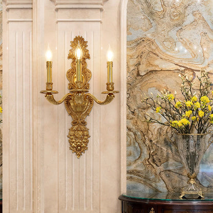 The luxury brass wall lamp installed on a light stone-textured wall beside a marble vase with yellow flowers.