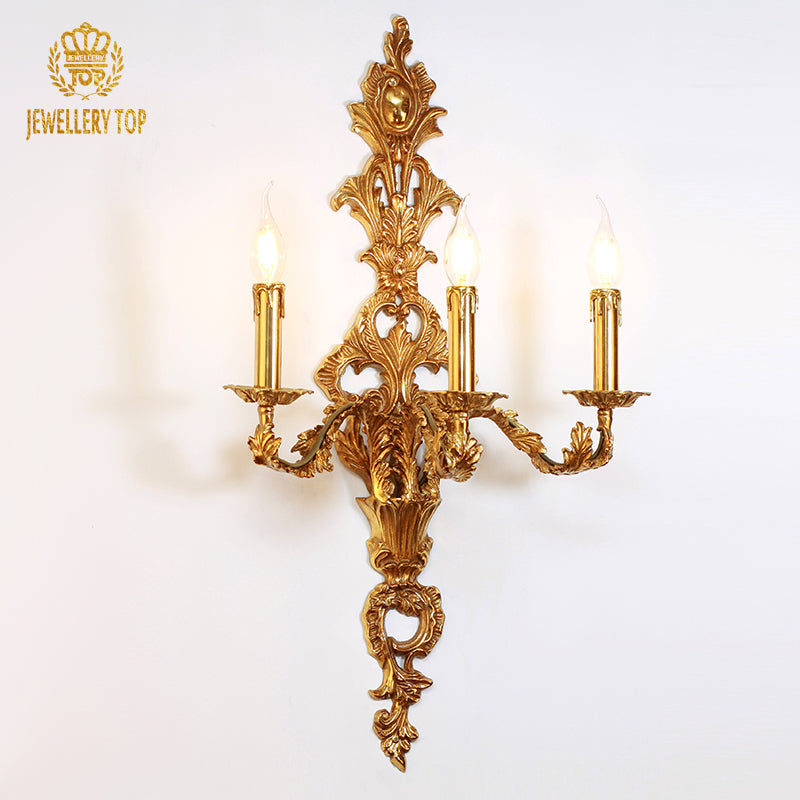 Branded close-up (JEWELLERY TOP) of the golden decorative wall lamp with three lit candle-style LED bulbs.