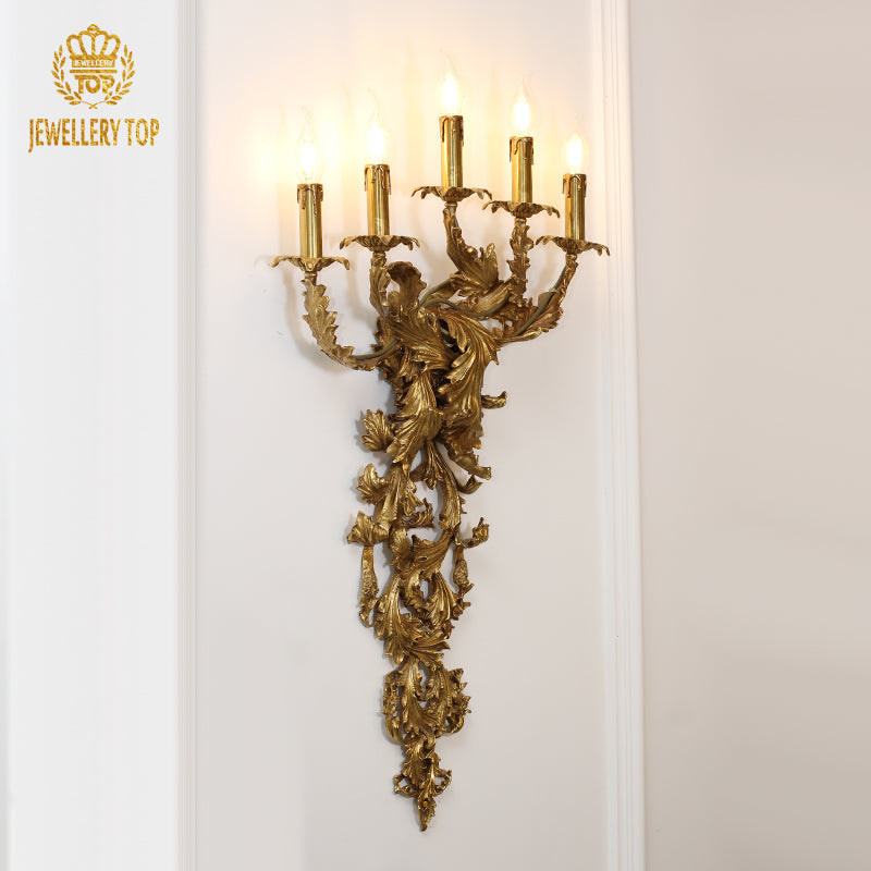 Branded product shot (JEWELLERY TOP) of the ornate five-candle brass sconce with intricate leaf carvings.