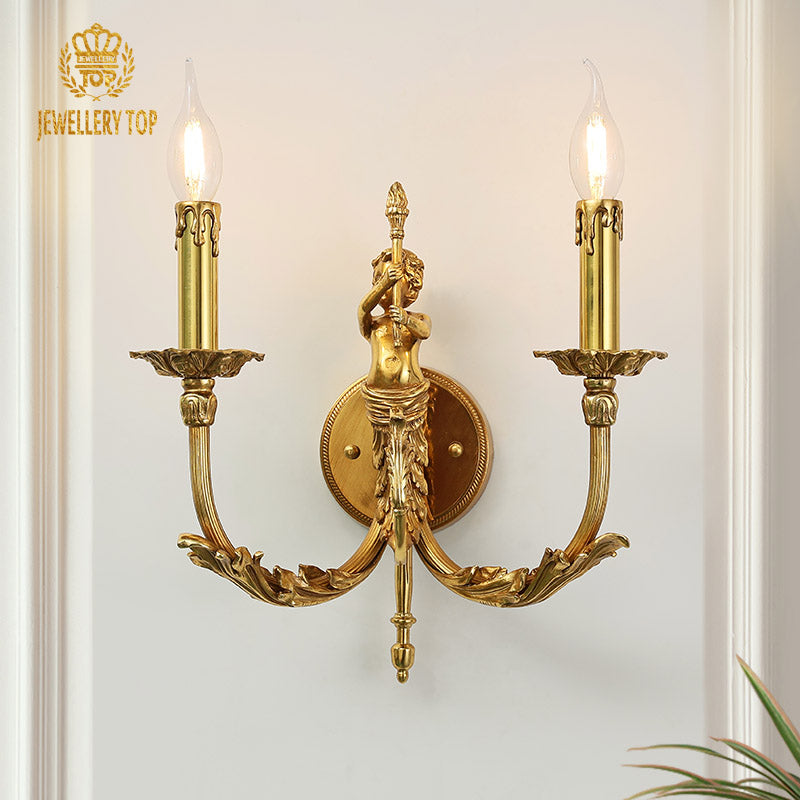 Eros Vigil Brass Wall Sconce with a detailed angel sculpture and two candle bulbs, mounted on a light wall with a green plant.