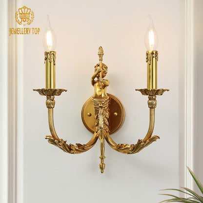 Eros Vigil Brass Wall Sconce with a detailed angel sculpture and two candle bulbs, mounted on a light wall with a green plant.