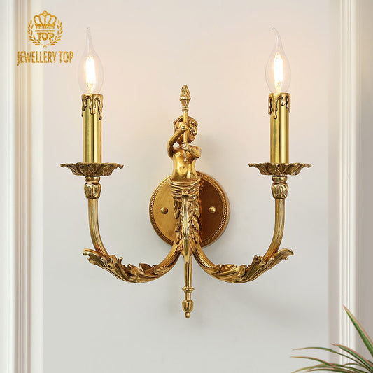 Eros Vigil Brass Wall Sconce with a detailed angel sculpture and two candle bulbs, mounted on a light wall with a green plant.