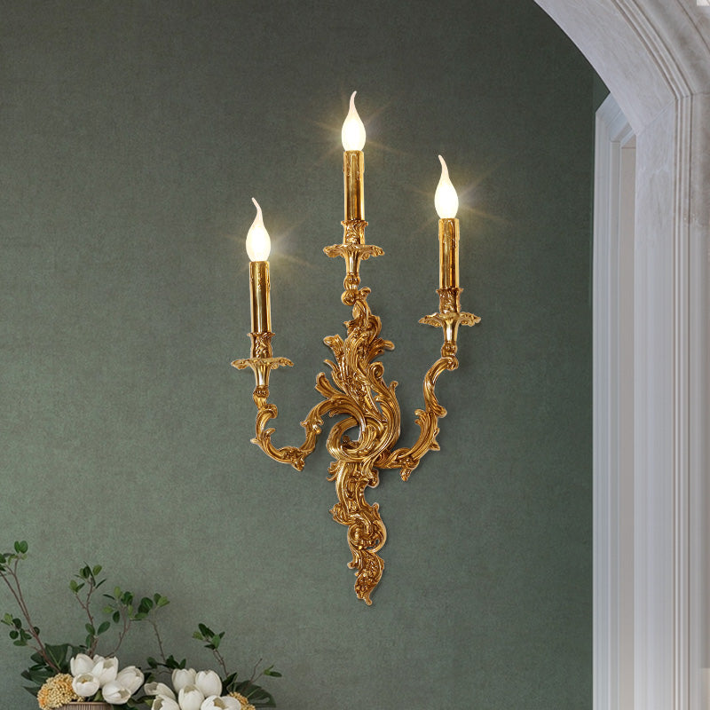 The installed classical brass candelabra sconce on a sage green wall, emitting a warm glow beside white trim and fresh lilies.